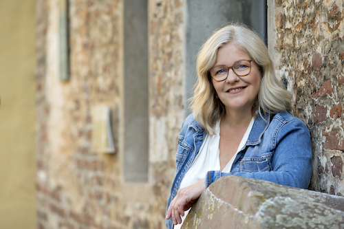 Ursula Proebsting – lange, silberne Haare, Jeansjacke – lehnt an einer Mauer aus alten Backsteinen. Sie lächelt in die Kamera.