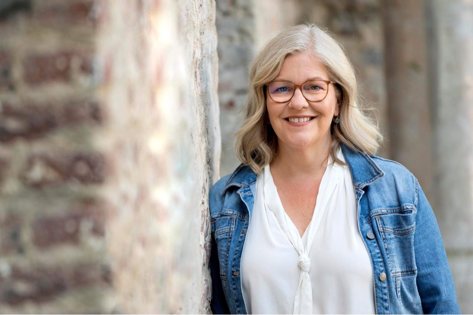 Ursula Proebsting steht an einer alten Bruchsteinmauer. Sie hat silberne, halblange Haare, trägt eine Jeansjacke und lächelt.
