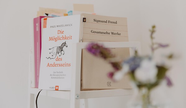 Ein Bücherstapel mit einer unscharfen Blumenvase im Vordergrund. Die Titel zweier Bücher sind zu lesen: „Die Möglichkeit anders zu sein“ von Paul Watzlawick und „Gesammelte Werke“ von Sigmund Freud.