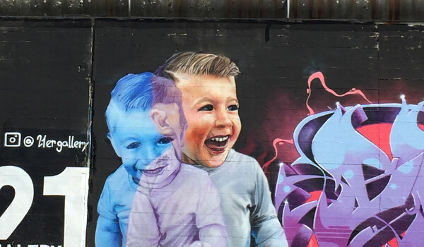 Eine Mauer mit einer Collage darauf mit zwei Kindern. Eines schaut nach unten und lächelt leicht, es ist bläulich eingefärbt und leicht durchsichtig. Halb dahinter ein etwas größeres Kind, das offen lacht und leicht nacht rechts schaut. Rechts neben den Kindern ist ein abstraktes Graffiti zu sehen. Links daneben zwei weisse Ziffern und etwas Handschrift, die aber nicht zu lesen ist.