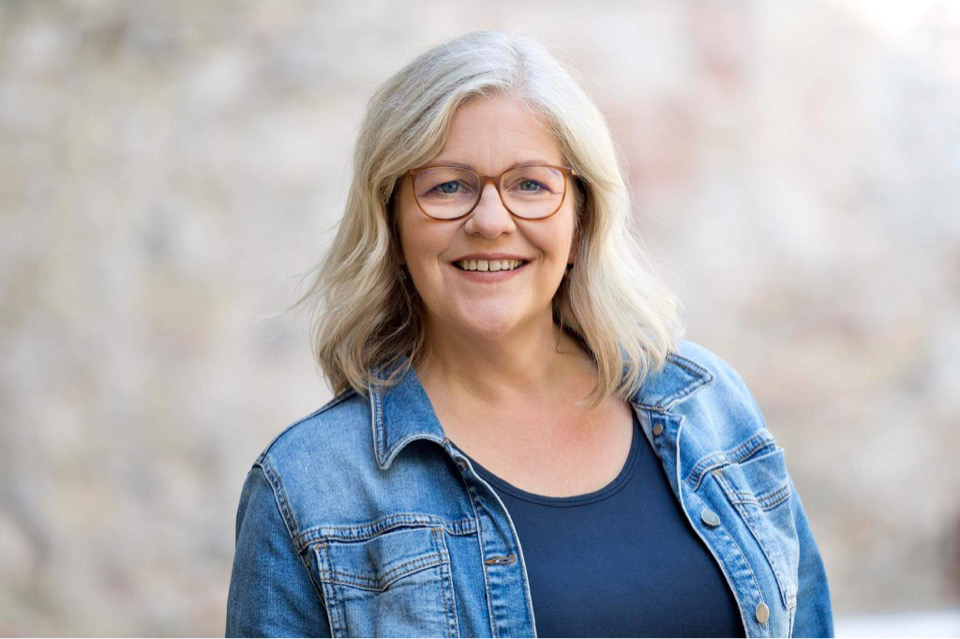 Ursula Proebsting – lange silberne Haare, Jeansjacke und Brille – schaut lächelnd in die Kamera.